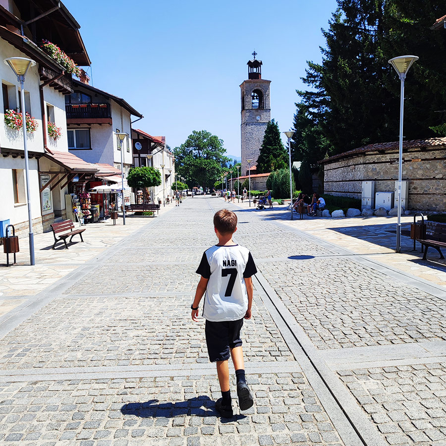 Child walking along the cobbled street of Bansko old town with Holy Trinity Church tower in the background, Bulgaria.