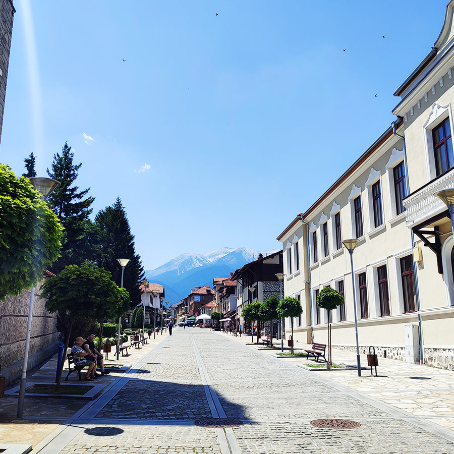 Main pedestrian street in Bansko, Bulgaria, with Pirin Mountain peaks visible in the distance.
