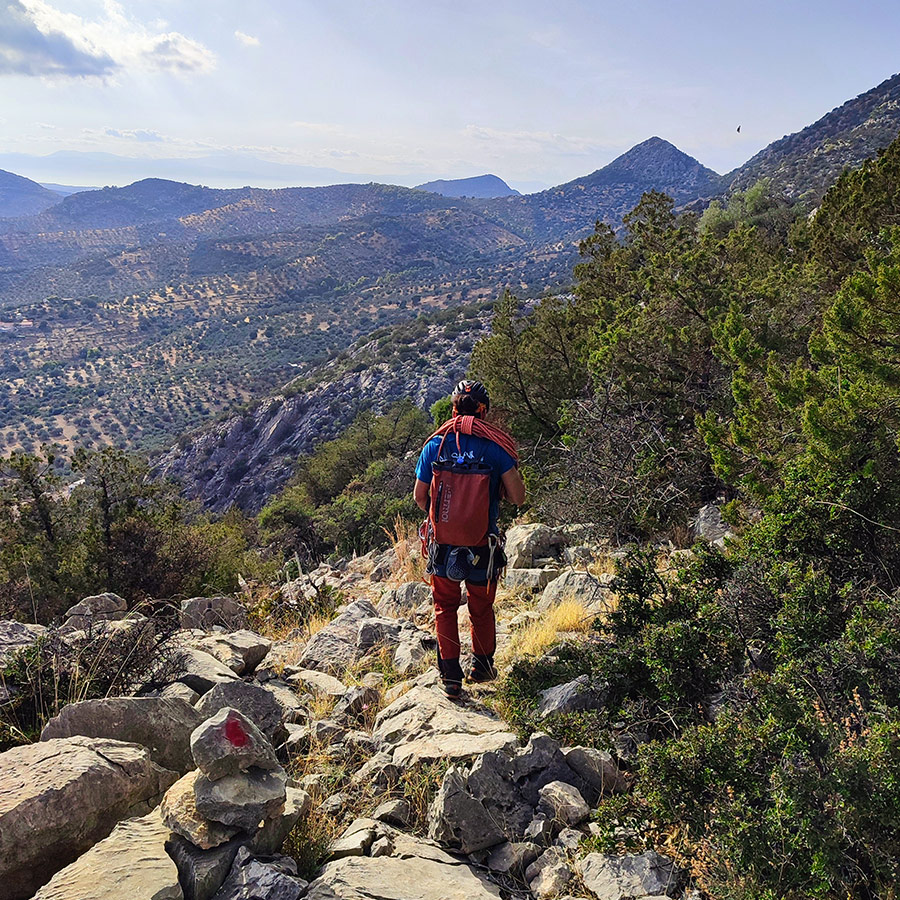 Climber hiking down rocky slope from Pleiades Crag with olive groves below.